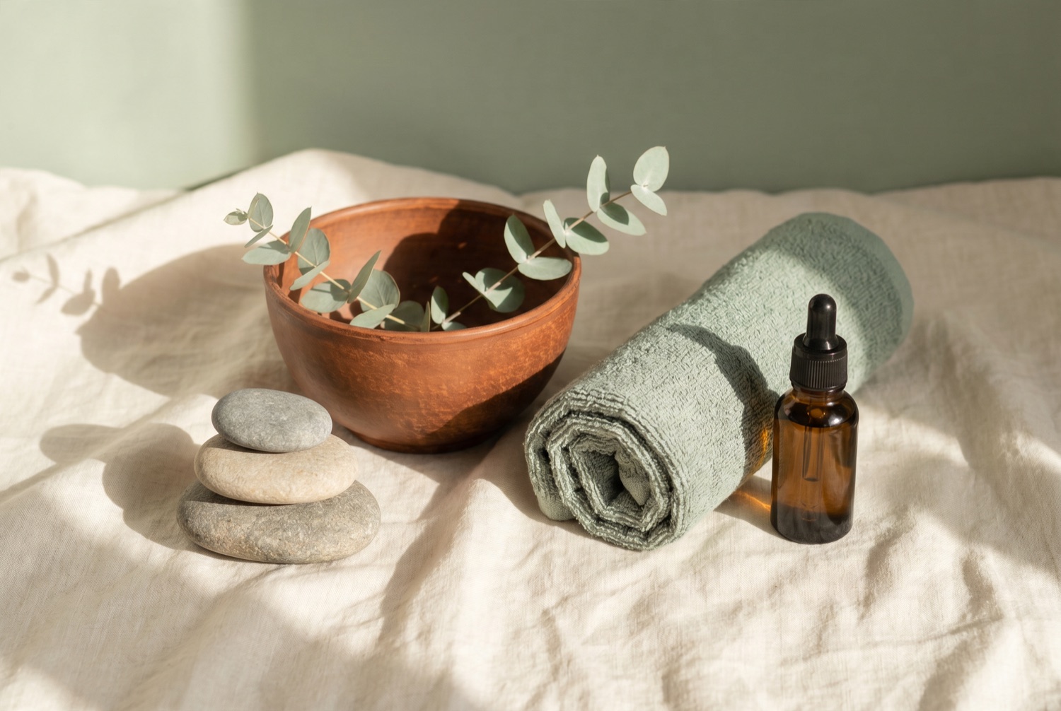 A calm still life of smooth stones, a clay bowl, eucalyptus and a rolled sage towel on cream linen.